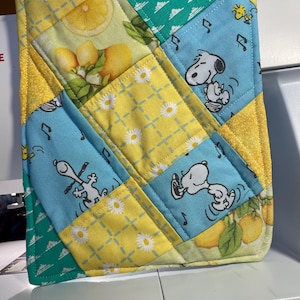 May include: A square quilted potholder featuring a patchwork design with alternating fabric patterns. The fabric includes light blue sections with Snoopy and Woodstock, yellow sections with white daisies, and green sections with lemons and leaves.