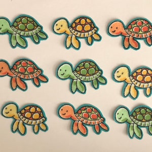 May include: Nine colorful felt turtle appliques with different colored shells and faces. The turtles are all facing the right side of the image.