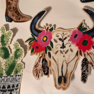 May include: A white bull skull with floral accents and feathers. The skull is painted with pink, red, and yellow flowers. The feathers are black and white.