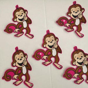 Puede incluir: Cinco parches de mono con cuerpos marrones, caras color canela y acentos rosas. Cada mono sonríe y tiene una cola rosa. Los parches están dispuestos sobre una superficie blanca, con una regla visible en el lado izquierdo.