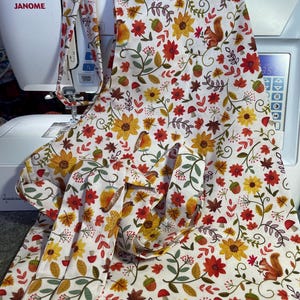 May include: A white apron featuring a repeating autumn-themed pattern of yellow sunflowers, red flowers, brown squirrels, small birds, green leaves, acorns, and red mushrooms. The apron includes long fabric ties and a neck loop for securing the garment.