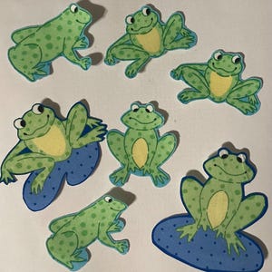 May include: Seven green frog appliques with yellow bellies and black eyes. Some frogs are sitting on blue lily pads with white polka dots.