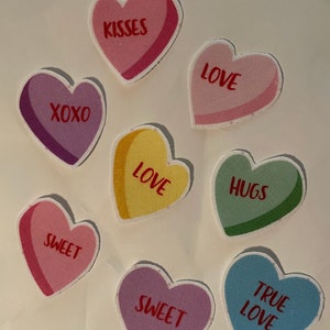 May include: Eight heart-shaped felt patches with different Valentine's Day messages. The patches are in various colors, including pink, purple, yellow, green, and blue. The messages on the patches include "XOXO", "Kisses", "Love", "Hugs", "Sweet", "True Love", and "Sweet".