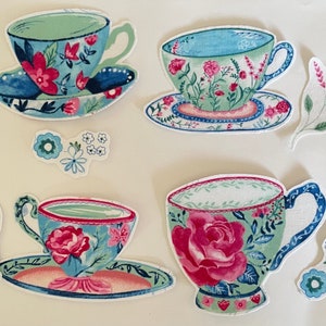 Op de afbeelding: Vier theekopjes en schoteltjes met bloemenpatronen in tinten blauw, groen, roze en wit. De theekopjes en schoteltjes zijn versierd met delicate bloemenontwerpen en hebben een vintage esthetiek.