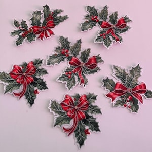 Può includere: Un set di sei applicazioni in tessuto ricamato raffiguranti foglie di agrifoglio verde scuro con bacche rosse e un fiocco di nastro rosso brillante. Ogni pezzo ha un bordo bianco e una trama cucita dettagliata.