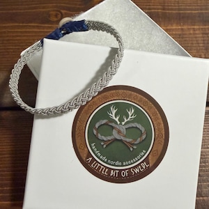May include: A silver-coloured braided wire bracelet with a blue leather closure and a light-coloured button fastener. The bracelet rests on a white square gift box featuring a circular sticker with an antler logo and the text A LITTLE BIT OF SWEDE.