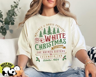 May include: Cream-colored t-shirt featuring a vintage-style design for "White Christmas." The graphic includes text, Christmas trees, and holly. The text reads "Wallace & Davis Present White Christmas Featuring The Haynes Sisters."