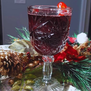 Può includere: Un bicchiere di vino rosso con una ciliegia come guarnizione, su un tavolo con un ramo di pino e ornamenti dorati.