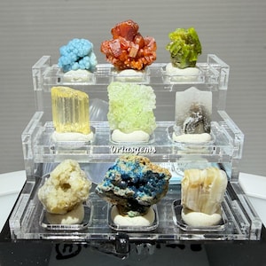 Könnte beinhalten: Ein dreistufiges Display aus klarem Acryl, das neun farbenfrohe Mineralien zeigt. Die Mineralien sind in Blau, Orange, Grün, Gelb und Weiß gehalten. Der Text "Ortasgems" ist auf dem Display sichtbar.