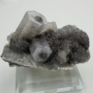 May include: A cluster of gray and white crystals, with a mix of sharp, geometric shapes and textured, layered formations. The crystals are set on a clear, rectangular display stand. The image is well-lit, highlighting the natural textures and colors.