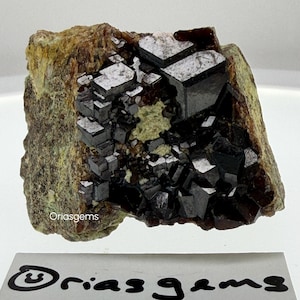 Op de afbeelding: Een natuurlijk mineraal specimen met donkere, glanzende, kubische kristallen gegroepeerd op een matrix van aarde bruin en groen gesteente. De kristallen hebben een reflecterend, metallic uiterlijk. De tekst "Oriasgems" is zichtbaar.
