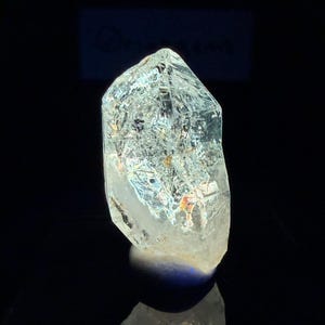 May include: A clear quartz crystal with a pointed tip. The crystal has internal fractures and inclusions, with iridescent colors visible within. The crystal is set against a black background, creating a contrast that highlights its clarity and form.