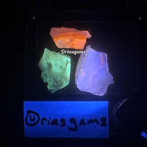 Op de afbeelding: Drie gloeiende mineraalmonsters in oranje, groene en paarse tinten onder zwart licht. De tekst "Oriasgems" is zichtbaar aan de boven- en onderkant van de afbeelding.