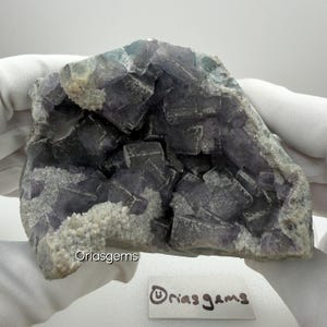 May include: A raw, natural fluorite specimen with a mix of purple, gray, and white colors. The fluorite features cubic crystal formations and a rough, textured surface. The image includes a small white tag with the text "Oriasgems".