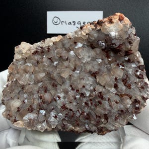 May include: A close-up of a mineral specimen with a crystalline structure. The mineral is primarily a translucent, light beige color, with clusters of reddish-brown inclusions scattered throughout. The surface has a rough, textured appearance. A small white tag with the text "@riasgems" is visible.