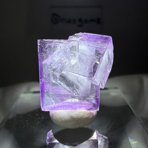 May include: A translucent, purple fluorite crystal with a cube-like structure. The crystal sits atop a small, rounded, white base. The image is well-lit, highlighting the crystal's facets and internal structures. The background is dark, emphasizing the crystal's form.