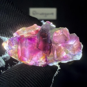 Op de afbeelding: Een ruw, veelzijdig kristalcluster met levendige tinten dieppaars, magenta en iriserend goud. Het mineraalexemplaar vertoont scherpe, hoekige punten en doorschijnende oppervlakken die het licht opvangen en een gloeiend effect creëren.