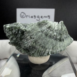 May include: A raw, uncut green mineral specimen with a fibrous texture. The stone is a mix of light and dark green hues, resting on a small, round, off-white base. The text "Oriasgems" is visible on a white card in the background.