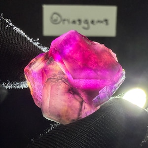 Op de afbeelding: Een doorschijnend, ruw edelsteenkristal met levendige tinten magenta, paars en geel. Het mineraal heeft een gefacetteerde, onregelmatige geometrische structuur met zichtbare interne breuken. Een witte kaart met de tekst "Oriasgems" is zichtbaar.
