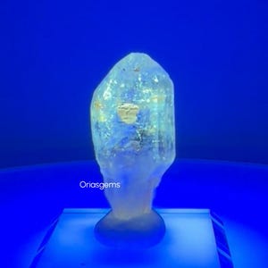 May include: A large, translucent crystal with a light blue hue, resting on a white platform against a deep blue background. The crystal has a rough, natural texture and a unique shape. The text "Oriasgems" is visible on the lower left.