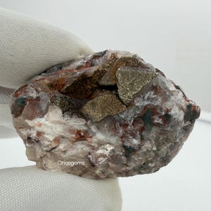 Può includere: Un esemplare minerale naturale grezzo con un mix di colori. La pietra presenta macchie di minerali metallici color oro, inclusioni rosse e marroni e aree di cristallo bianco e trasparente. La consistenza è ruvida e irregolare, con una varietà di forme e dimensioni.