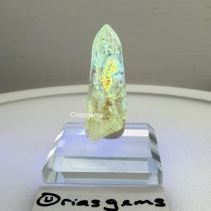 May include: A translucent, light green crystal with internal yellow and blue iridescent patterns. The crystal is displayed on a clear acrylic stand. The text "Oriasgems" is visible on the crystal and the stand.
