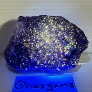 May include: A large, irregularly shaped, dark purple mineral specimen with a speckled, glowing yellow and green surface. The mineral sits on a blue base with the text "Oriasgems".