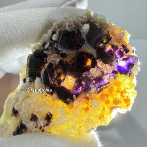 May include: A close-up of a fluorite mineral specimen. The fluorite crystals are a deep purple and black color, with a yellow and white base. The image is well-lit, showcasing the mineral's natural beauty. The text "Oriasgems" is visible.