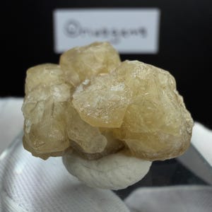 May include: A cluster of pale yellow mineral crystals, possibly a type of calcite or quartz, sits atop a small, rounded, off-white base. The crystals have a rough, textured surface and a translucent quality, with visible internal structures. The background is dark.