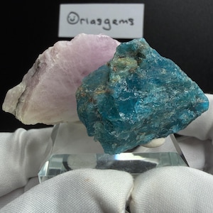 May include: A close-up of two raw mineral specimens. One is a light pink color, and the other is a vibrant blue. The minerals are displayed on a clear, angled stand. The text "Oriasgems" is visible in the background.