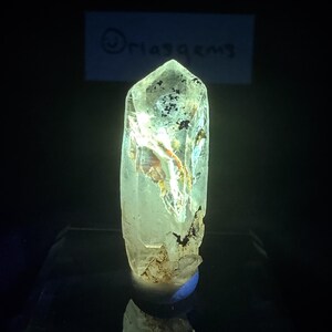 May include: A translucent, elongated quartz crystal point with a pointed termination. The crystal features internal fractures, light-reflecting facets, and dark mineral inclusions. A rectangular sign with the text "Jriaggems" is visible behind it.