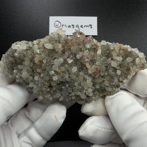 Pode incluir: Um aglomerado de cristais de quartzo com tons de roxo e castanho. Os cristais são segurados por mãos enluvadas contra um fundo preto. O texto "Oriasgems" é visível em um rótulo branco.
