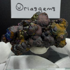 May include: A cluster of iridescent mineral specimens with a rough, textured surface. The colours range from deep purple and brown to vibrant yellows, greens, and blues. The text "Oriasgems" is written on a white card in the background.
