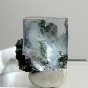 以下が含まれることがあります： 淡い紫色の縁と緑色のインクルージョンを持つ、透明な立方体型の蛍石の結晶。結晶は、濃い緑色の鉱物ベースと白く丸みを帯びた石の上にあります。鉱物標本です。