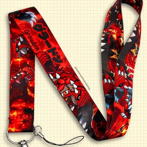 Könnte beinhalten: Rotes Schlüsselband mit einem sich wiederholenden Muster eines roten und schwarzen Monsters mit weißen Zähnen und Krallen. Auf dem Schlüsselband stehen das Wort "Groudon" und japanische Schriftzeichen. Es enthält einen silbernen Metallverschluss und eine schwarze Schnur.