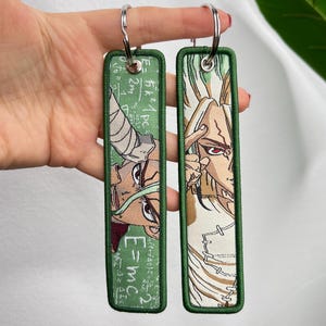 Può includere: Due portachiavi rettangolari verdi con disegni di personaggi anime ricamati. I portachiavi hanno un bordo verde e anelli argentati. Un portachiavi presenta l'equazione E=mc². L'altro portachiavi ha un diverso design di personaggi anime.