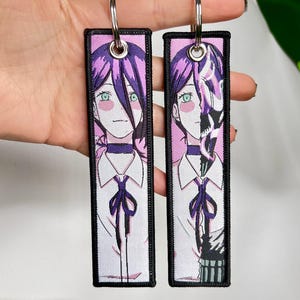 Puede incluir: Dos llaveros rectangulares con arte de personajes de estilo anime. Cada llavero presenta un retrato de una persona con cabello morado, ojos verdes y una camisa blanca con una corbata morada. Los llaveros tienen bordes negros y llaveros plateados.