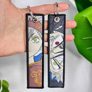 Puede incluir: Dos llaveros rectangulares con diseños de personajes de anime. Uno presenta un personaje con cabello blanco y ojos verdes, el otro con cabello negro y ojos rojos. Ambos tienen un borde negro y un llavero plateado.