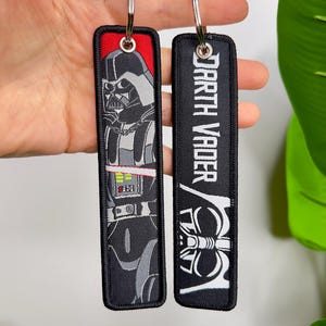 Puede incluir: Dos llaveros rectangulares con aros plateados. Uno presenta a Darth Vader en gris y negro sobre un fondo rojo. El otro llavero es negro con las palabras "Darth Vader" en blanco y un casco blanco de Darth Vader.