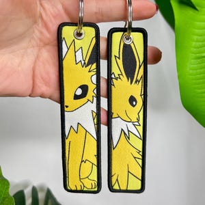 May include: Two yellow and black embroidered keychains featuring a cartoon character with a white collar and black accents. The keychains have silver metal rings at the top for attaching to keys or bags. The background is a light colour.