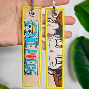 Puede incluir: Dos llaveros rectangulares con bordes amarillos y anillas plateadas. Un llavero presenta un logotipo de calavera y huesos cruzados con la palabra "ONE PIECE" en azul y texto japonés. El otro llavero tiene una ilustración de personaje.
