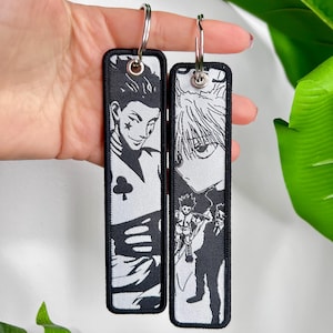 Op de afbeelding: Twee rechthoekige zwart-witte sleutelhangers met anime-personages. De sleutelhangers hebben een zwarte rand en een zilveren metalen ring. Eén sleutelhanger toont een personage met een speelkaartsymbool, de andere toont een personage met stekelig haar.