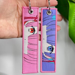 Op de afbeelding: Twee rechthoekige sleutelhangers met anime-ogen. Eén sleutelhanger heeft roze haar en een rood oog, de andere heeft paars haar en een blauw oog. Beide hebben een roze rand en een zilveren sleutelring.