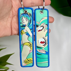 Puede incluir: Dos llaveros bordados con un personaje de dibujos animados con pelo azul y un bikini amarillo y negro. Los llaveros tienen un fondo turquesa con un borde azul y anillas plateadas.