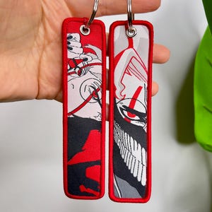 Può includere: Due portachiavi rettangolari con bordi rossi e anelli argentati. Ogni portachiavi presenta un design unico in rosso, nero, bianco e grigio. Un design raffigura una figura con contorno rosso, l'altro un volto stilizzato.