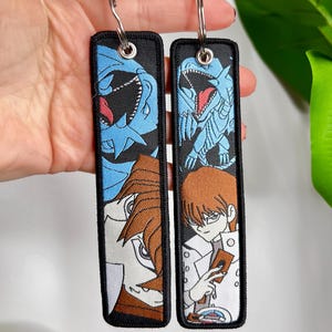 May include: Two black rectangular keychains with embroidered anime-style character designs. One features a blue dragon and a character with brown hair. The other shows a character in a white coat holding a card. Each keychain has a silver keyring.