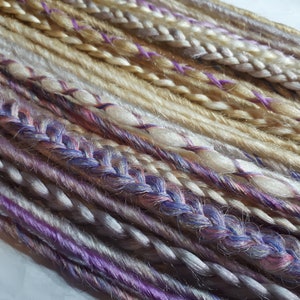 Puede incluir: Una colección de extensiones de cabello sintético en varios tonos de rubio, blanco y morado. Las extensiones están trenzadas y tienen un aspecto texturizado.