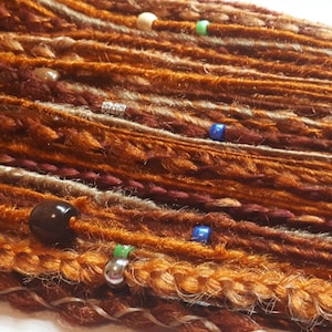 May include: A close-up of a set of brown, blonde, and burgundy synthetic dreadlocks with beads and metal rings. The dreadlocks are arranged in a row and are ready to be installed.