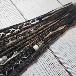 May include: Close-up of dark grey and brown dreadlocks with decorative white and silver accents. The dreadlocks are adorned with beads and wrapped sections, creating a textured and stylish appearance. The hair extensions are arranged on a light wood surface.