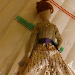 May include: A doll wearing a gold and white dress with a purple sash. The dress has a sparkly, textured fabric and gold leaf accents. The doll has red hair and is wearing a brown jacket.
